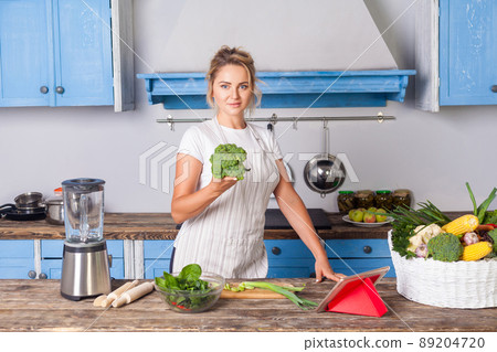 Happy attractive woman in apron holding broccoli and smiling at camera, cooking salad in modern kitchen, preparing vegetarian food, basket of fresh vegetables on table, vegan diet healthy nutrition Happy attractive woman in apron holding broccoli and smiling at camera, cooking salad in modern kitchen, preparing vegetarian food, basket of fresh vegetables on table, vegan diet healthy nutrition 89204720