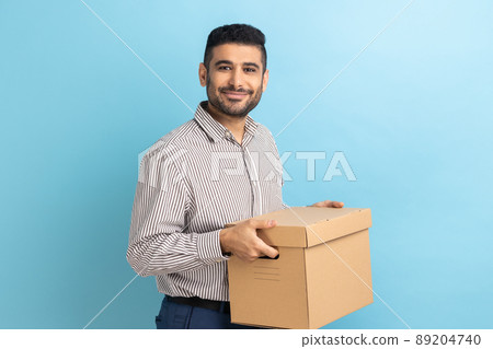 Smiling positive businessman holding cardboard parcel, delivering orders to address, looking at camera with friendly expression, wearing striped shirt. Indoor studio shot isolated on blue background. 89204740