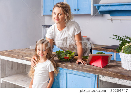 Cute woman and her daughter smiling at camera standing in modern kitchen, mother keeping hand on girl's shoulder, cooked salad with fresh green vegetables on table, vegetarian food, healthy nutrition Cute woman and her daughter smiling at camera standing in modern kitchen, mother keeping hand on girl's shoulder, cooked salad with fresh green vegetables on table, vegetarian food, healthy nutrition 89204747