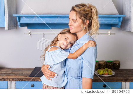 Beautiful mother hugging lovely daughter and looking at camera with toothy smile, standing in kitchen with modern blue furniture and enjoying together. indoor, home family togetherness. 89204763