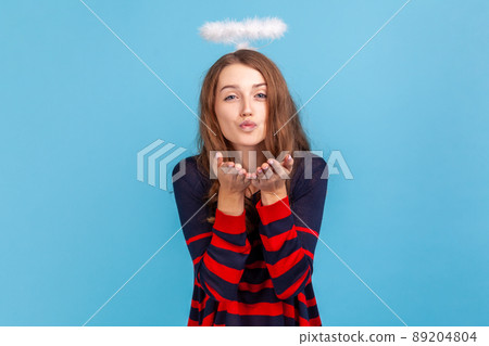 Kind woman wearing striped casual style sweater and nimb over head, sending air kiss over palms, expressing fondness, romantic feelings. Indoor studio shot isolated on blue background. 89204804