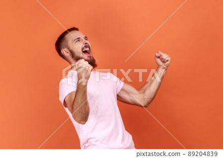 Portrait of overjoyed handsome bearded man expressing winning gesture with raised fists and screaming, celebrating victory, wearing pink T-shirt. Indoor studio shot isolated on orange background. Portrait of overjoyed handsome bearded man expressing winning gesture with raised fists and screaming, celebrating victory, wearing pink T-shirt. Indoor studio shot isolated on orange background. 89204839
