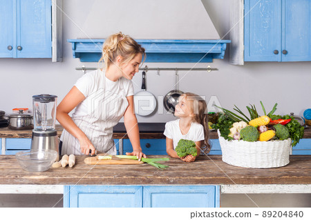 Happy pretty mother in apron chopping cutting green vegetable with knife and smiling to little daughter, woman cooking salad in kitchen, preparing vegetarian food, basket of fresh vegetables on table Happy pretty mother in apron chopping cutting green vegetable with knife and smiling to little daughter, woman cooking salad in kitchen, preparing vegetarian food, basket of fresh vegetables on table 89204840