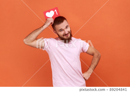 Portrait of bearded man holding like icon above head, looking at camera with satisfied facial expression, social media, wearing pink T-shirt. Indoor studio shot isolated on orange background. Portrait of bearded man holding like icon above head, looking at camera with satisfied facial expression, social media, wearing pink T-shirt. Indoor studio shot isolated on orange background. 89204841