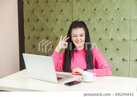 Portrait of positive funny young woman with black dreadlocks hairstyle in pink blouse sitting in cafe and showing ok sign with hand, winking, looking at camera and toothy smile. Indoor, lifestyle Portrait of positive funny young woman with black dreadlocks hairstyle in pink blouse sitting in cafe and showing ok sign with hand, winking, looking at camera and toothy smile. Indoor, lifestyle 89204842
