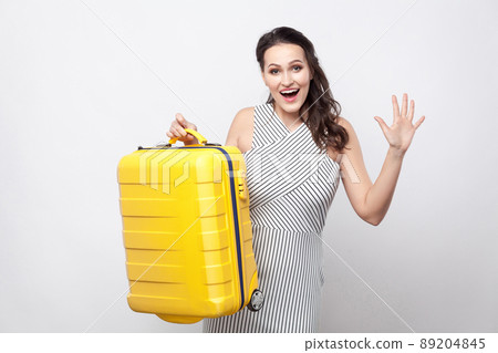 Portrait of happy beautiful young traveller in striped dress standing and holding yellow suitcase greeting toothy smile and ready for vacation. Indoor studio shot,isolated,copy space, grey background 89204845