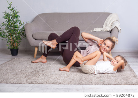 Young cheerful woman doing crunches looking at camera, mother working on abs together with her preschool daughter, happy family doing gymnastics at home, fitness training with child. body care, health 89204872