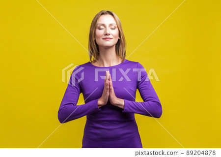 Harmony of mind. Portrait of elegant woman in purple dress standing with closed eyes and peaceful calm face meditating, holding hands in prayer. indoor studio shot isolated on yellow background Harmony of mind. Portrait of elegant woman in purple dress standing with closed eyes and peaceful calm face meditating, holding hands in prayer. indoor studio shot isolated on yellow background 89204878