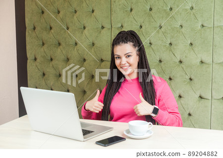 Portrait of attractive positive young girl freelancer with black dreadlocks hairstyle in pink blouse sitting in cafe and working on laptop with toothy smile and showing thumb up, looking at camera. 89204882