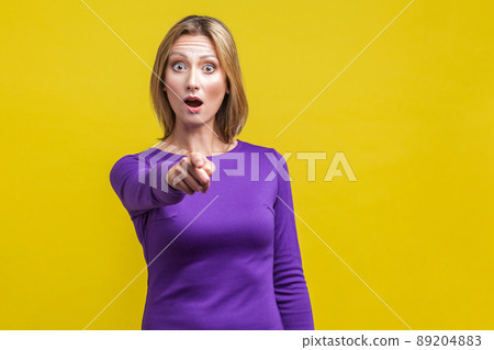 Portrait of wondered woman in tight purple dress pointing at camera, looking shocked amazed with open mouth, hard to believe. indoor studio shot isolated on yellow background 89204883