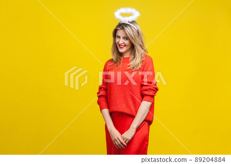 Portrait of modest angel beautiful young woman with bright makeup in red casual clothes standing with halo on her head and looking shy at camera. indoor studio shot isolated on yellow background 89204884