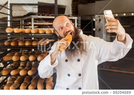 Portrait of hungry young adult blogger baker with long beard in white uniform standing in factory and making selfie on shelves with fresh bread background, tasty loaf, Indoor, profession concept 89204889