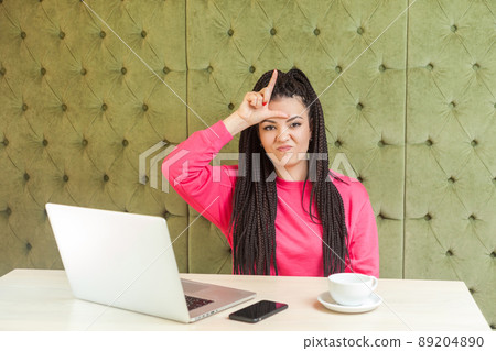 Negative rude young girl with black dreadlocks hairstyle in pink blouse is sitting in cafe and having bad mood, showing loser sing with finger and looking to camera. indoor 89204890