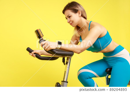 Side view of athletic woman riding exercise bike and looking tutorial video on smart phone, cardio workout, wearing blue sportswear. Indoor studio shot isolated on yellow background. Side view of athletic woman riding exercise bike and looking tutorial video on smart phone, cardio workout, wearing blue sportswear. Indoor studio shot isolated on yellow background. 89204893