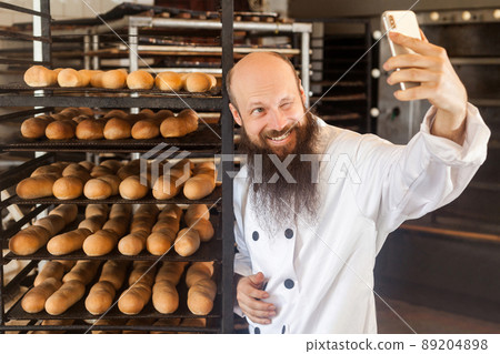 Portrait of cheerful young adult blogger baker with long beard in white uniform standing in factory and making selfie on shelves with fresh bread background, winking, Indoor, profession concept 89204898