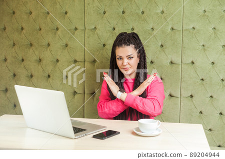 Warning aggressive young girl with black dreadlocks in pink blouse are sitting in cafe and working on laptop, showing crossing raised arms like stop or end gesture, looking at camera. Indoor 89204944
