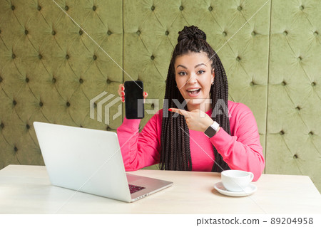 Portrait of beautiful amazed young blogger with dreadlocks hairstyle in pink blouse is sitting, holding mobile phone and pointing finger, looking at camera with shoked face. Indoor, business style Portrait of beautiful amazed young blogger with dreadlocks hairstyle in pink blouse is sitting, holding mobile phone and pointing finger, looking at camera with shoked face. Indoor, business style 89204958