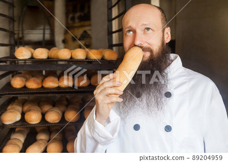 Portrait of cheerful young adult baker with long beard in white uniform standing in his manufacture, smelling with pleasure fresh bread and enjoying. looking up, indoor, profession concept Portrait of cheerful young adult baker with long beard in white uniform standing in his manufacture, smelling with pleasure fresh bread and enjoying. looking up, indoor, profession concept 89204959