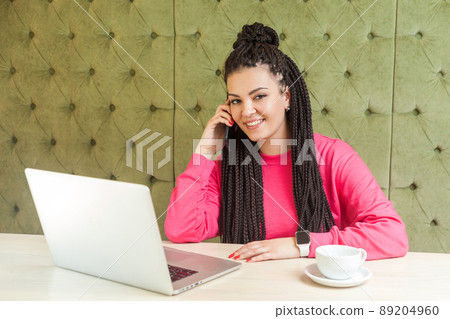 Portrait of beautiful satisfied happy young businesswoman with black dreadlock hairstyle in pink blouse sitting in cafe, talking on phone and toothy smile, looking at camera. Indoor, healthy lifestyle Portrait of beautiful satisfied happy young businesswoman with black dreadlock hairstyle in pink blouse sitting in cafe, talking on phone and toothy smile, looking at camera. Indoor, healthy lifestyle 89204960