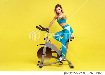 Full length portrait of blonde woman coach on riding bike simulator, pointing to camera, choosing you for personal workout, wearing blue sportswear. Indoor studio shot isolated on yellow background. 89204965