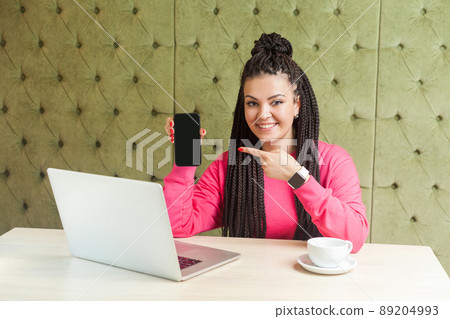 Portrait of beautiful satisfied happy young businesswoman with black dreadlocks in pink blouse is sitting in cafe, holding phone and pointing finger, looking at camera with toothy smile. Indoor Portrait of beautiful satisfied happy young businesswoman with black dreadlocks in pink blouse is sitting in cafe, holding phone and pointing finger, looking at camera with toothy smile. Indoor 89204993