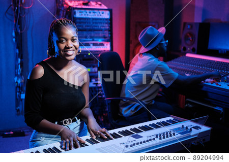 Joyful young African American female musician playing digital keyboard while producer working on mixing console in recording studio 89204994
