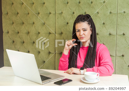 Portrait of attractive young girl with black dreadlocks hairstyle in pink blouse are sitting in cafe, working and showing little size with finger, looking at camera. Indoor, lifestyle 89204997