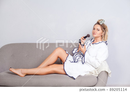 Positive attractive young woman with curlers in hair in robe holding face powder and looking at camera with smile, using cosmetics, doing make up on sofa at home, beauty and fashion concept, indoor 89205114