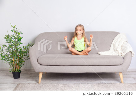 Cute calm little girl meditating in lotus pose with closed eyes sitting on couch at home, child doing mudra gesture, peaceful happy face expression, having yoga practice, relaxing dreaming indoor 89205116