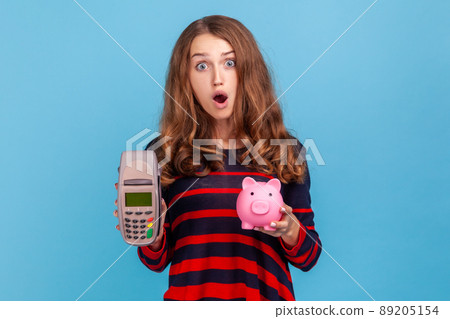 Astonished woman wearing striped casual style sweater, showing pos payment terminal and empty screen smartphone, has surprised expression. Indoor studio shot isolated on blue background. 89205154
