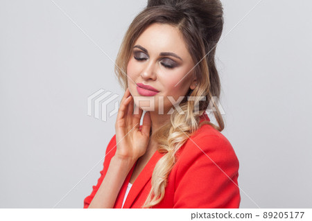 Portrait of beautiful business lady with hairstyle and makeup in red fancy blazer standing and touching her face and smiling with closed eyes. indoor studio shot, isolated on grey background. Portrait of beautiful business lady with hairstyle and makeup in red fancy blazer standing and touching her face and smiling with closed eyes. indoor studio shot, isolated on grey background. 89205177