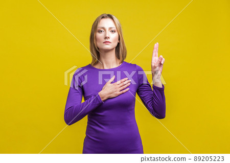 Portrait of honest citizen, young woman in elegant tight purple dress standing with hand on chest and fingers up, making loyalty promise. indoor studio shot isolated on yellow background 89205223