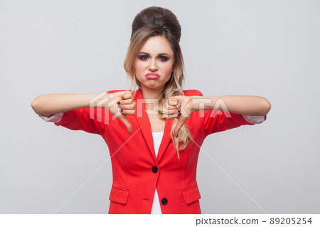 Portrait of sad dissatisfied beautiful business lady with hairstyle and makeup in red fancy blazer, standing with thubms down and looking at camera. indoor studio shot, isolated on grey background. 89205254