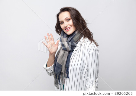 Portrait of happy beautiful brunette woman in white striped jacket and blue scarf standing, looking at camera with toothy smile and greeting. indoor studio shot, isolated on light grey background. 89205265
