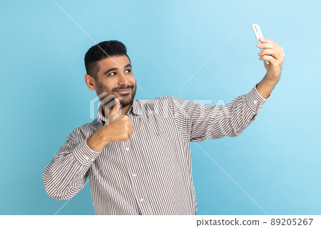 Satisfied man blogger showing thumbs up like gesture and winking looking at phone camera, making selfie or recording video, wearing striped shirt. Indoor studio shot isolated on blue background. 89205267