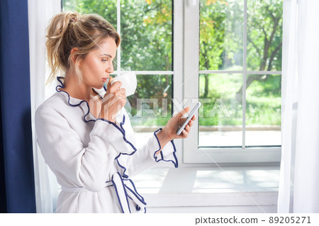 Young busy attractive woman in robe with beautiful hairstyle standing at window holding mobile phone and drinking coffee, reading news or message in morning, using smartphone with serious expression 89205271
