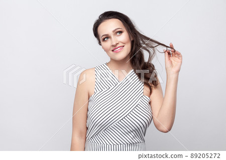 Happy optimistic beautiful young brunette woman with makeup and striped dress standing touching hair, dreaming and looking away with toothy smile. indoor studio shot, isolated on grey background. 89205272
