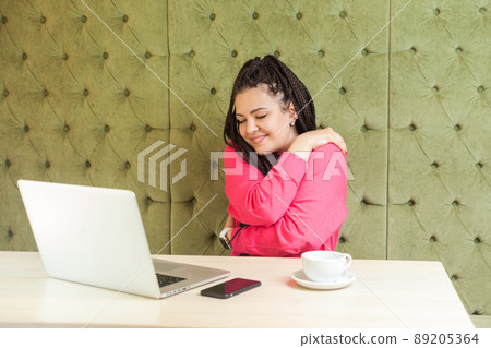 Portrait of lovely romantic young girl freelancer with black dreadlocks hairstyle in pink blouse are sitting alone in cafe and hugging self with closed eyes and toothy smile. Indoor, lifestyle Portrait of lovely romantic young girl freelancer with black dreadlocks hairstyle in pink blouse are sitting alone in cafe and hugging self with closed eyes and toothy smile. Indoor, lifestyle 89205364