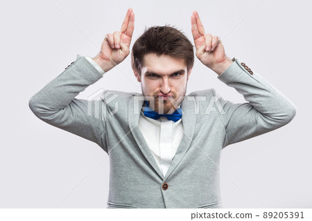 Portrait of playful handsome bearded man in casual grey suit and blue bow tie standing and looking at camera with funny face and horns fingers. indoor studio shot, isolated on light grey background. 89205391