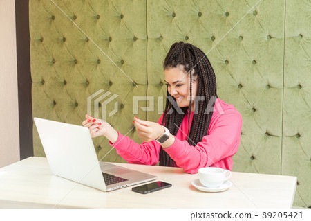 Interesting young freelancer with black dreadlocks hairstyle in pink blouse are sitting in cafe and agrees with his employer for payment through webcam and showing cash gesture with happy face. Indoor 89205421