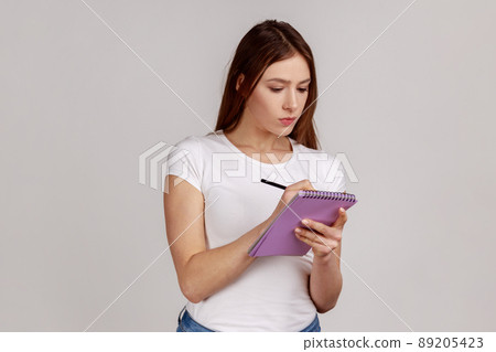 Portrait of clever serious woman writing in notebook, completing to-do list or studying, having concentrated expression, wearing white T-shirt. Indoor studio shot isolated on gray background. Portrait of clever serious woman writing in notebook, completing to-do list or studying, having concentrated expression, wearing white T-shirt. Indoor studio shot isolated on gray background. 89205423
