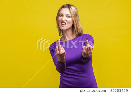 Portrait of impolite emotional woman in elegant tight purple dress standing showing middle finger at camera, looking with hate and disrespect grimace. indoor studio shot isolated on yellow background 89205439
