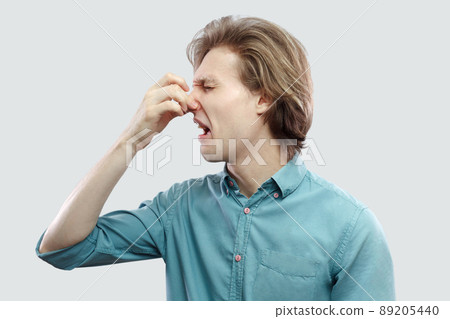 profile side view portrait of unhappy handsome long haired blonde young man in blue casual shirt standing pinching his nose because bad smell. indoor studio shot, isolated on light grey background. 89205440