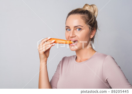 Portrait of young woman biting orange carrot, fresh raw vegetables, concept of healthy eating with vitamins beta carotene, vegetarian diet, low calorie food. studio shot isolated on grey background 89205445