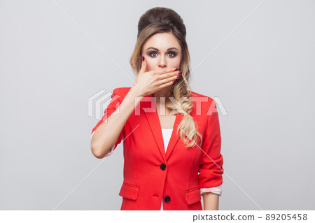 Portrait of shocked beautiful business lady with hairstyle and makeup in red fancy blazer, standing and covering her mouth and looking at camera with big eyes. studio shot, isolated on grey background 89205458