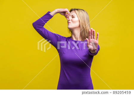 Unpleasant smell. Portrait of young woman in purple dress pinching her nose with fingers to hold breath, frowning in displeasure and showing stop gesture. studio shot isolated on yellow background Unpleasant smell. Portrait of young woman in purple dress pinching her nose with fingers to hold breath, frowning in displeasure and showing stop gesture. studio shot isolated on yellow background 89205459