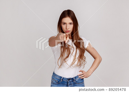 Portrait of strict displeased young woman making choice with indicating finger, looking bossy and dissatisfied, showing guilty, wearing white T-shirt. Indoor studio shot isolated on gray background. 89205478