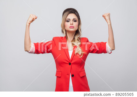 I am strong. Portrait of proud beautiful business lady with hairstyle and makeup in red fancy blazer, standing with raised arms and looking at camera. indoor studio shot, isolated on grey background. 89205505