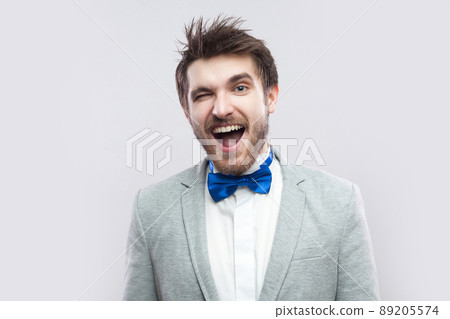 Portrait of funny handsome bearded man in casual grey suit and blue bow tie standing looking at camera and winking. indoor studio shot, isolated on light grey background. 89205574