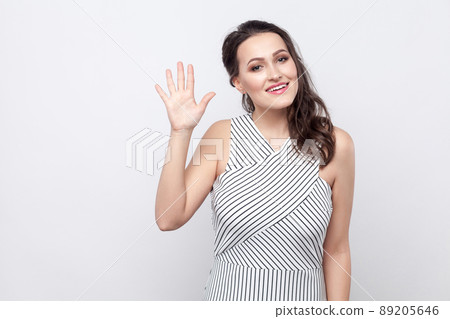 Portrait of beautiful happy young brunette woman with makeup and striped dress standing, looking at camera with toothy smile and greeting gesture. indoor studio shot, isolated on grey background. 89205646